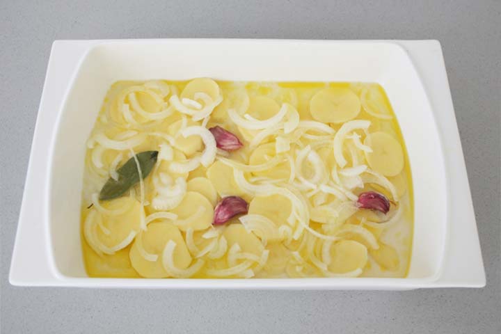 Rodajas de patata y cebolla en una fuente junto a unos dientes de ajo y una hoja de laurel